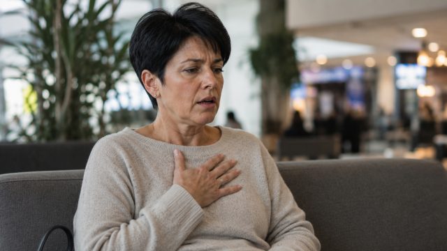 Vrouw van rond de 55 jaar met kort zwart haar die gespannen hoog op de borst ademt op een bank in een openbare ruimte